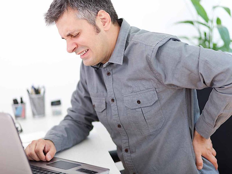 Das Bild zeigt einen Mann sitzend vor einem Laptop mit Rückenschmerzen.