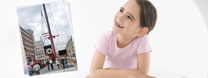 Das Bild zeigt ein kleines Mädchen wie es glücklich guckt mit einem Foto neben ihr. Das Foto zeigt wie ein neues MRT-Gerät geliefert wird.